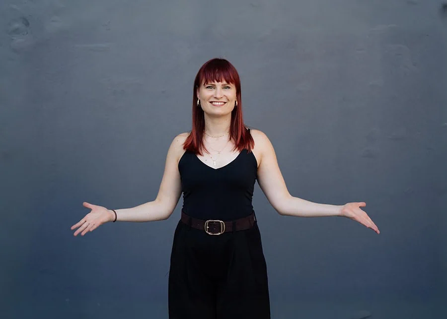 Mareike Walter steht mit ausgebreiteten Armen vor einem grauen Hintergrund und trägt ein schwarzes Oberteil und eine schwarze Hose.