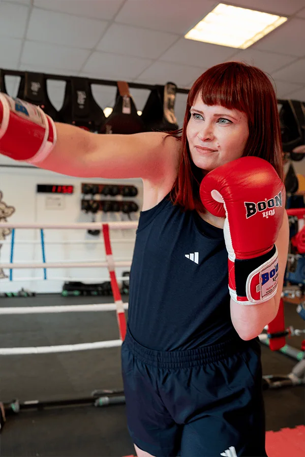 Eine Boxerin mit roten Boxhandschuhen, die einen Jab im Training ausführt, in einem Boxring mit Trainingsausrüstung im Hintergrund.