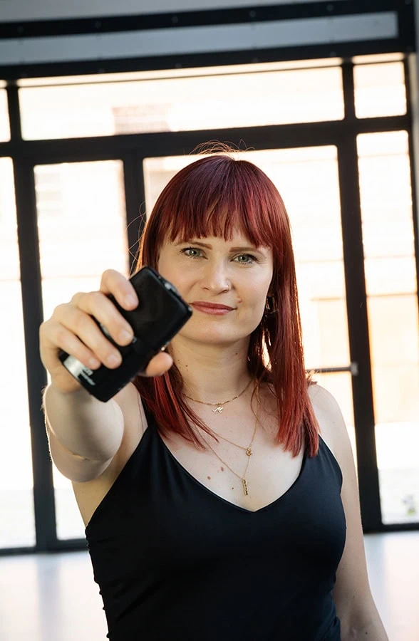 Frau mit rotem Haar in schwarzem Oberteil, die ein Mikrofon in der Hand hält und in die Kamera blickt.