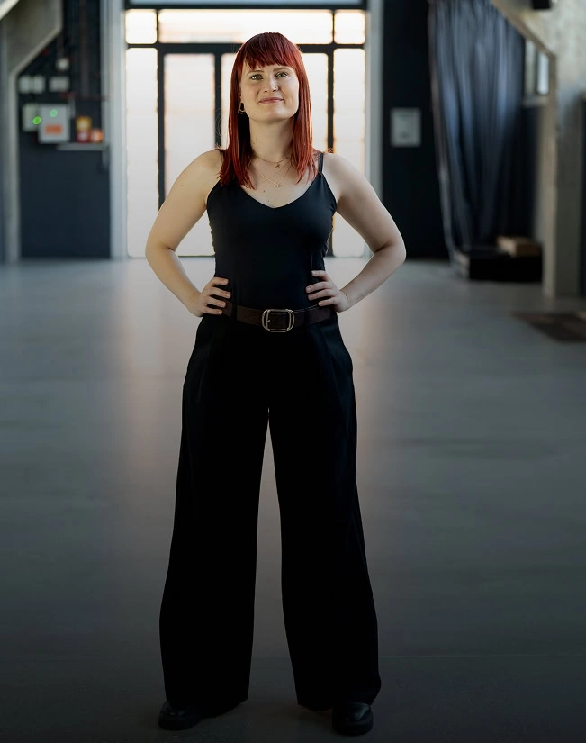 Mareike Walter steht selbstbewusst in einem schwarzen Outfit mit rotem Haar und einem Gürtel in einem modernen Raum.